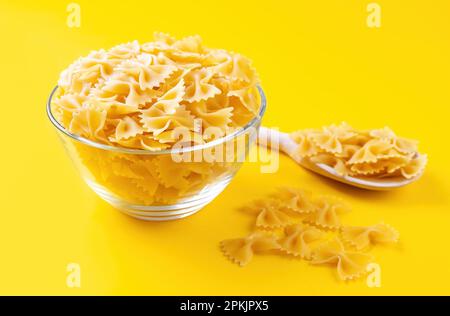 Bowl and wooden spoon with pasta on yellow background. Raw pasta farfalle, butterfly shape, ingredient for cook, traditonal italian cuisine Stock Photo