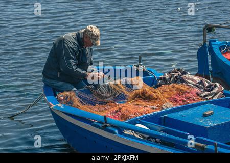 Fisherman with a hat repairing with a big needle a fishing net on an old blue wooden fishing boat at the port quay in Bari, Puglia Italy Stock Photo