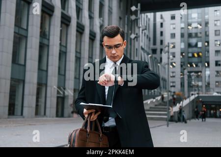 With notepad in hands. Businessman in black suit and tie is outdoors in the city Stock Photo