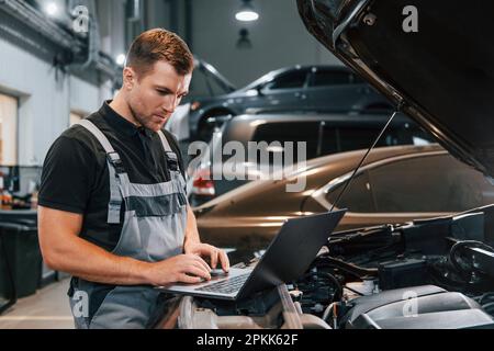 Using laptop. Man in uniform is working in the auto service Stock Photo