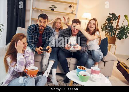 Playing video game. Group of friends have party indoors together. Stock Photo