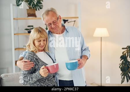 Standing and holding cups of drinks. Senior man and woman is together at home. Stock Photo