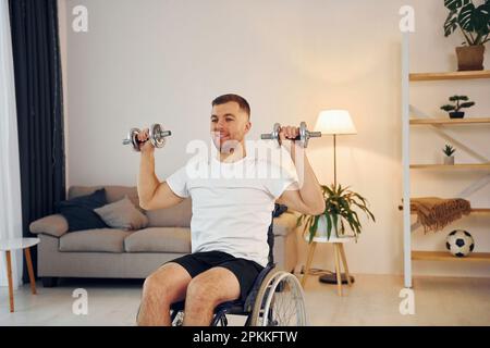 Doing exercises by using dumbbells. Disabled man in wheelchair is at home. Stock Photo