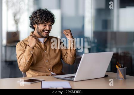 Successful hispanic businessman programmer celebrating victory and triumph, man inside office sitting at table and working using laptop, holding hands up gesture of achieving good results. Stock Photo
