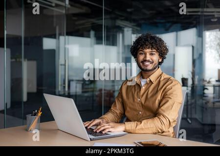 Portrait of young successful Indian man at workplace inside office, businessman smiling and looking at camera, man at work using laptop, programmer with curly hair coding software. Stock Photo