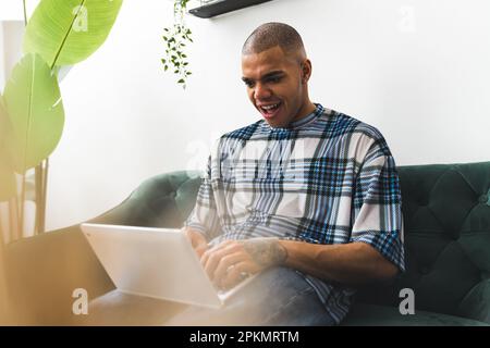 young Afro-American tattooed man looking at something unexpecting on a laptop while sitting on a sofa at home. High quality photo Stock Photo