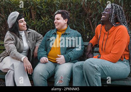Three diverse young friends, a brunette woman, a non-binary plus size person, and an African woman with dreadlocks, casually dressed, sitting on a par Stock Photo