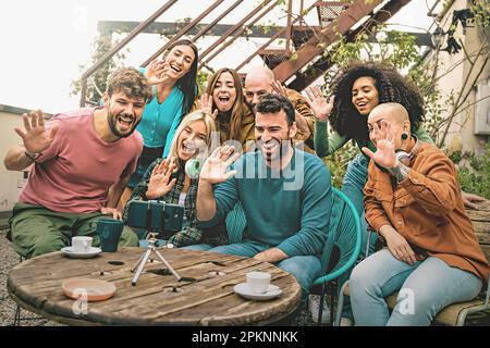 Seven diverse friends sitting around a wooden table on a terrace, smiling and waving at a smartphone on a tripod, having a video call. One is a non-bi Stock Photo