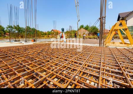 Pile of rusty rectangle steel reinforcement ready for installation placed at construction site. Workers in background are pouring concrete into buildi Stock Photo