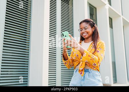 Young hispanic happy woman browsing on internet using a cellphone device. Latin teenage female texting messages on a social media using a smart phone app and wireless technology standing outdoors. High quality photo Stock Photo