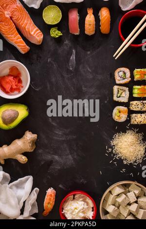 Overhead shot of sushi and ingredients on dark background. Sushi rolls, nigiri, raw salmon steak, rice, cream cheese, avocado, lime, pickled ginger (g Stock Photo