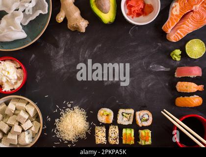 Overhead shot of sushi and ingredients on dark background. Sushi rolls, nigiri, raw salmon steak, rice, cream cheese, avocado, lime, pickled ginger (g Stock Photo