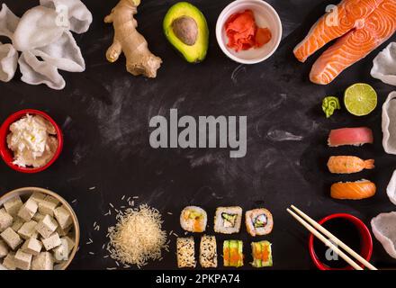 Overhead shot of sushi and ingredients on dark background. Sushi rolls, nigiri, raw salmon steak, rice, cream cheese, avocado, lime, pickled ginger (g Stock Photo