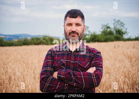 Arms crossed mature farmer man looking serious at camera standing at wheat field. Front view of folded agricultural rural worker with pensive attitude at harvest farm land. High quality photo Stock Photo