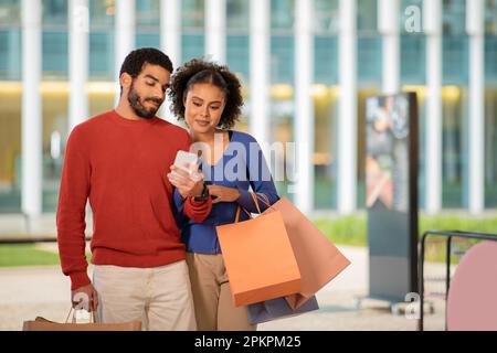 Happy Multicultural Couple Doing Shopping Using Cellphone Standing Outside Stock Photo