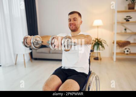 Doing exercises by using dumbbells. Disabled man in wheelchair is at home Stock Photo