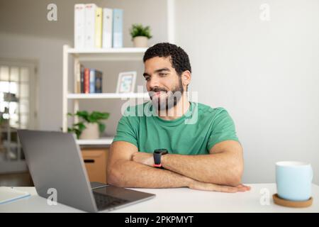 Smiling handsome millennial arabic man with beard has video call, working with laptop at workplace in room Stock Photo