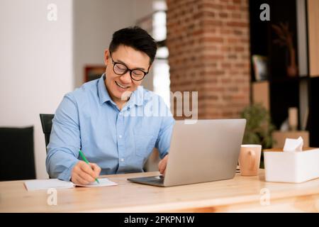 Happy asian man making notes, analyzing data for investment at workplace with laptop computer in home office interior Stock Photo