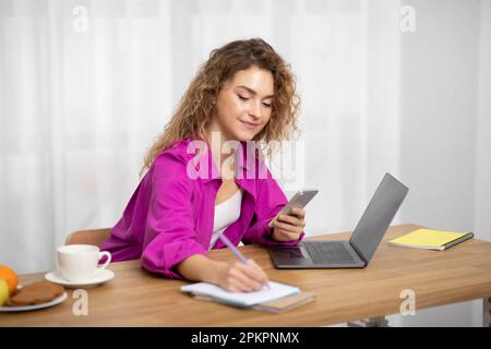 Beautiful Young Freelancer Woman Using Smartphone And Writing In Notepad Stock Photo