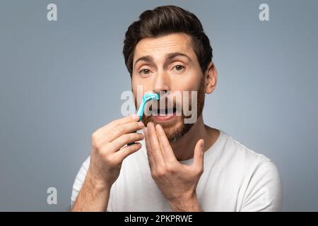 Handsome middle aged bearded man shaving mustache with razor and looking at camera over grey background Stock Photo