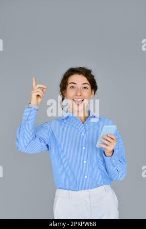 Young happy surprised business woman using phone pointing up isolated on grey. Stock Photo