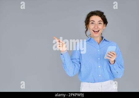 Young excited business woman using phone pointing aside isolated on grey. Stock Photo
