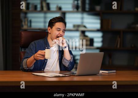 Sleepy employee sitting at dark office, using laptop, drinking coffee Stock Photo