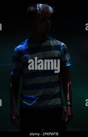 A man in a striped T-shirt stands in a dark room with a soft white and blue light falling on him Stock Photo