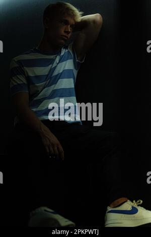 A thinking man in a striped T-shirt sits in a dark room with a soft natural light falling on him Stock Photo