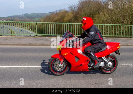 1999 90s nineties Red Honda Vfr800f V Four Motorcycle  Sports Tourer Petrol 781 cc with a rider wearing Red Mandeep Motorbike Helmet Cover Fluffy Funny plush full face helmets, crossing motorway bridge in Greater Manchester, UK Stock Photo