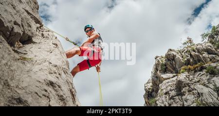 Active climber middle age man in protective helmet looking at camera while abseiling from cliff rock wall using rope with belay device and climbing ha Stock Photo