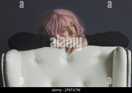 A portrait of a man in a pink wig, who is leaning on a white armchair, looks away. close-up. Stock Photo