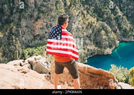 Young man standing on a rock cliff with US flag on his back and looking at sea beneath. Veteran traveller wearing American flag on his back while Stock Photo