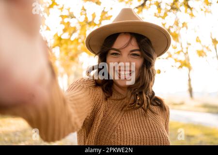 Smiling woman in trendy sweater having fun in autumn park. Adorable girl taking selfie on blur fall nature background. Copy space Stock Photo