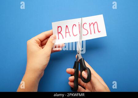 Hand holding card with the text racism. Top view of black scissors and cutted paper card with word Racism on the blue background. Top view Stock Photo