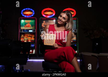 Portrait of a Young Beautiful and Well Dressed Caucasian Girl in Red Dress Playing in the Casino and Celebrate Holding Bingo Wood Game Card Stock Photo