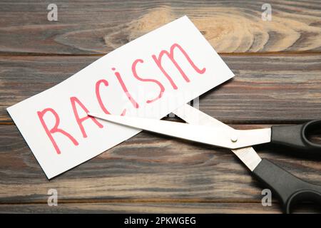 Top view of black scissors and cutted paper card with word Racism on brown background. Top view Stock Photo
