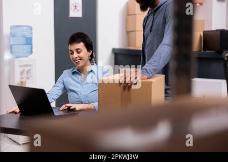 Storehouse workers looking at shipping logistics on laptop computer discussing cargo stock while working with carton boxes in warehouse. Distribution center fulfillment company Stock Photo
