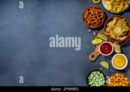 Various unhealthy salty bar snacks set. Dark grey table background with traditional party snacks - chips, onion rings, salted nuts, crisps,  pretzels Stock Photo
