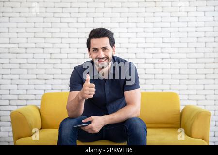 Lifestyle portrait of attractive handsome man with pleasant smile relaxing and sitting on yellow sofa at living room Stock Photo