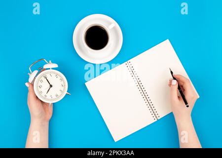 Female hands write in a notebook and hold an alarm clock, flat lay. Stock Photo