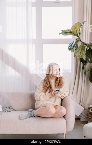 Thoughtful young woman in cozy sweater on couch looking out window Stock Photo