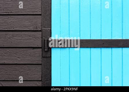 A two dimensional design created by a blue painted wooden door with a black hinge and dark wooden cladding Stock Photo