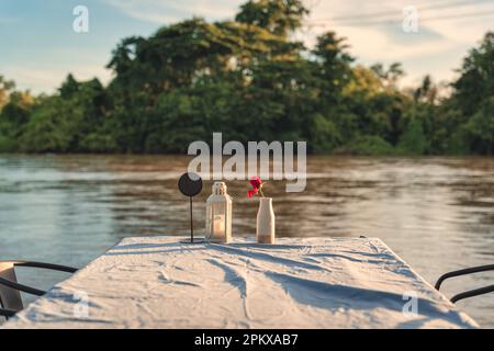 Flower vase, candle lantern, label on dining table with tablecloth at restaurant by riverfront in the sunset Stock Photo
