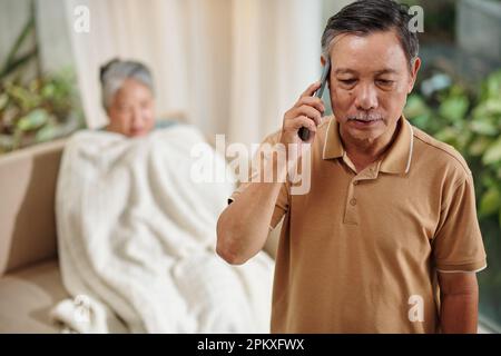 Serious senior man talking on phone, describing symptoms to doctor, his sick wife is sitting in background Stock Photo