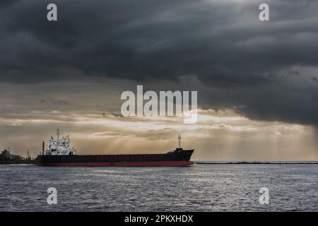 view of a tanker sailing across the sea under a dramatic dark sky with golden sunlight shining through the clouds. Stock Photo