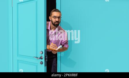Bearded man in glasses peeking out blue door, attentively looking and making notes. Inspector, accountant, journalist Stock Photo