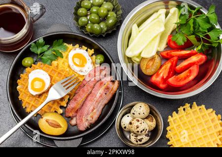 Chopped vegetables and herbs in a bowl. Bacon, waffles, fried eggs and a fork in a frying pan. Olives and quail eggs in bowls. A cup of tea. Flat lay. Stock Photo