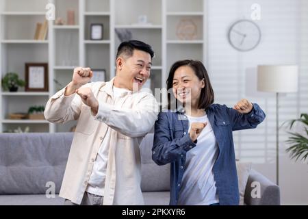 Happy Asian couple man and woman dancing together at home in daytime in living room, happy young family celebrating happy day and wedding anniversary. Stock Photo