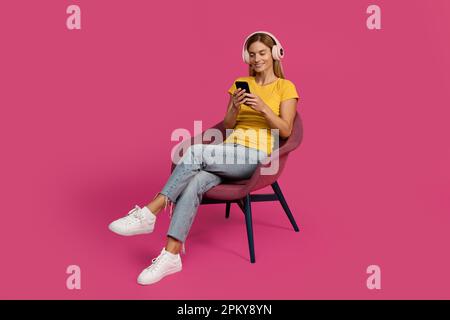 Happy middle aged european lady sit in armchair, typing on smartphone, listen music in wireless headphones Stock Photo